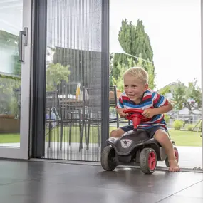 Ein Junge fährt lachend auf einem Bobby-Car durch eine offene Terrassentür mit Insektenschutz.