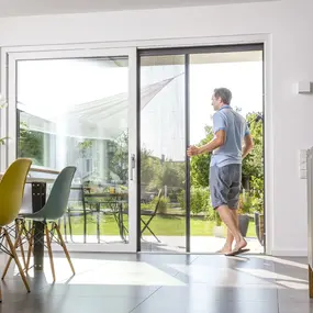 Mann öffnet eine große Terrassentür mit Insektenschutz im modernen, hellen Wohnzimmer.