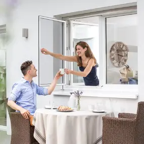 Frau reicht Mann Kaffee durch geöffnetes Insektenschutzfenster auf sonniger Terrasse.