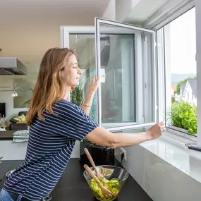 Frau lüftet die Küche beim Kochen, frische Luft und Kräuter am Fenster sorgen für Wohlbefinden.