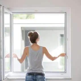 Junge Frau lüftet ein großes Fenster im modernen, hellen Wohnzimmer für frische Luft.