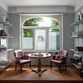 Gemütliche Leseecke mit zwei Sesseln, Bücherregalen, rundem Tisch und Gitarre am Fenster.