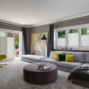 Modernes Wohnzimmer mit grauem Sofa, runden Couchtisch und großen Fenstern mit Plissees.