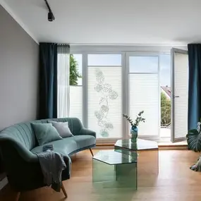 Modernes Wohnzimmer mit Glas-Couchtisch, grünem Sofa und großen bodentiefen Fenstern.