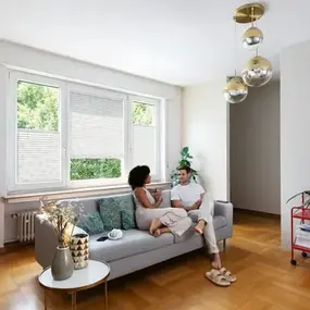 Paar sitzt entspannt auf Sofa im modernen Wohnzimmer mit großen Fenstern und Holzboden.