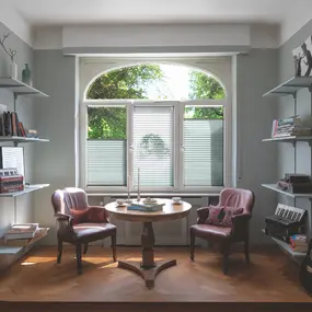 Helles Wohnzimmer mit großen Fenstern, Holzboden, Ledersesseln, Tisch und Bücherregalen.