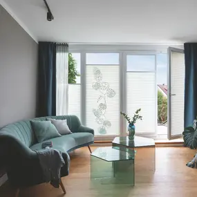 Modernes Wohnzimmer mit grüner Couch, Glas-Couchtisch und großen Fensterplissees.