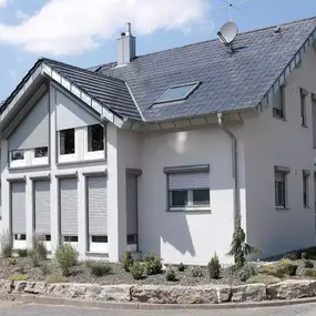 Modernes Einfamilienhaus mit Satteldach, Rollläden und gepflegtem Vorgarten bei Sonnenschein.