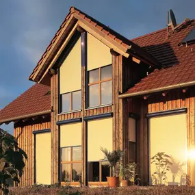 Modernes Holzhaus mit großen Fenstern, geschlossenen Rollläden und Solaranlage auf dem Dach.
