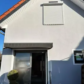 Einfamilienhaus mit weißer Fassade, Rollladen und moderner Haustür bei blauem Himmel.