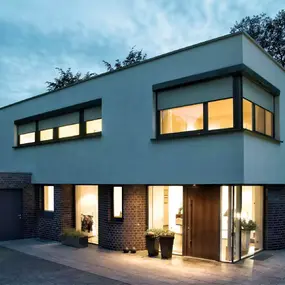 Modernes Einfamilienhaus mit Flachdach, großen Fenstern und beleuchtetem Eingangsbereich.