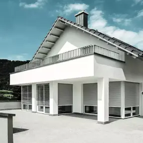 Modernes Einfamilienhaus mit Flachdach, Terrasse und Rollläden vor großen Fenstern.
