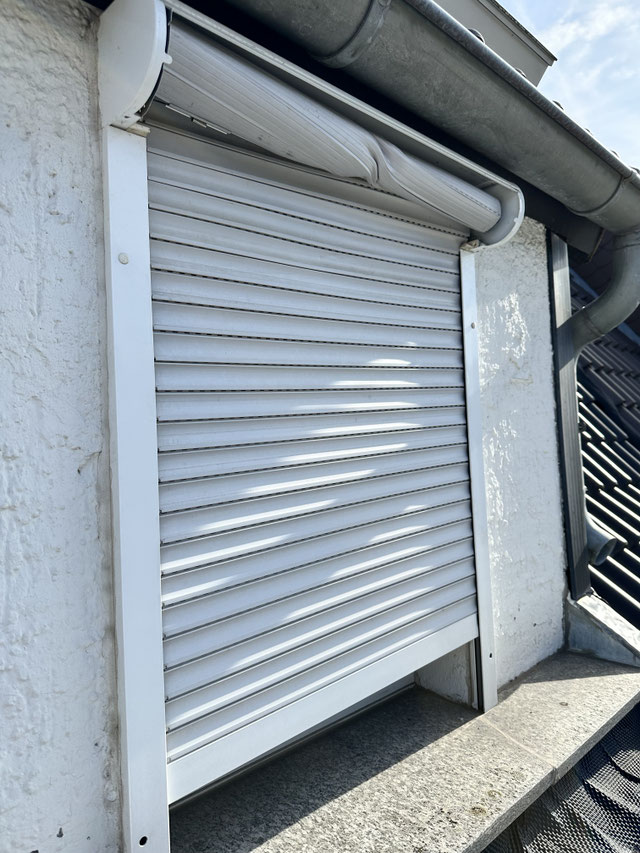 Beschädigter weißer Rollladen an einem Fenster unter einem Dach mit Regenrinne und Sonnenlicht.