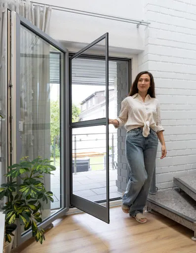 Frau öffnet moderne Glastür zur Terrasse in hellem Wohnraum mit Pflanze