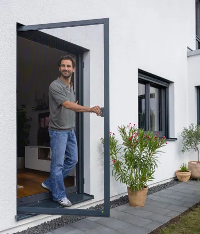 Mann öffnet moderne Terrassentür am weißen Haus, Gartenpflanzen daneben