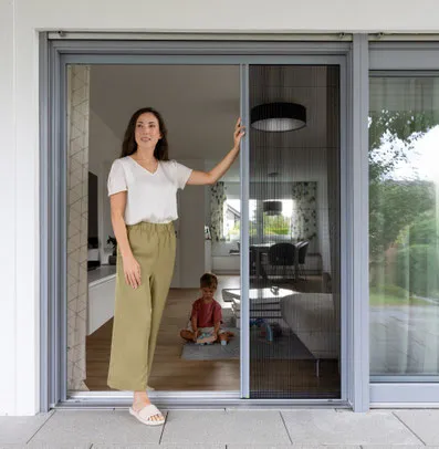 Frau öffnet Schiebetür mit Insektenschutz, Kind spielt im Wohnzimmer